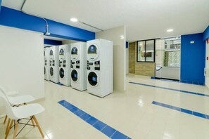 A laundry facility with multiple washing machines, a seating area, and a window with a view. - Apartahotel Livinnx Barranquilla (Puerto Colombia)