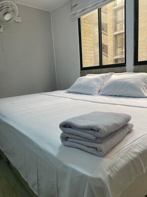 A neatly made bed with white linens and pillows, a standing fan, and a window with a view of a brick building. - Apartahotel Livinnx Barranquilla (Puerto Colombia)