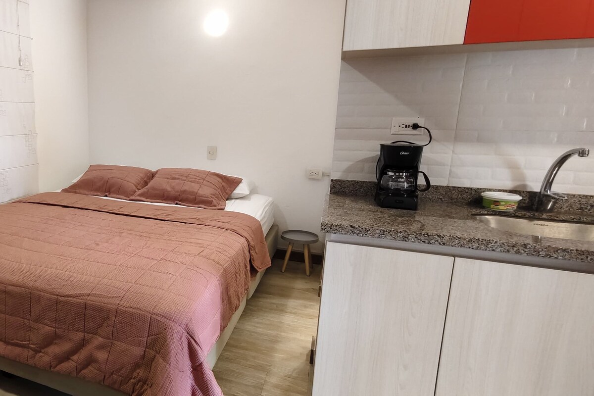 Un dormitorio con cama, un taburete pequeño y una kitchenette con cafetera.