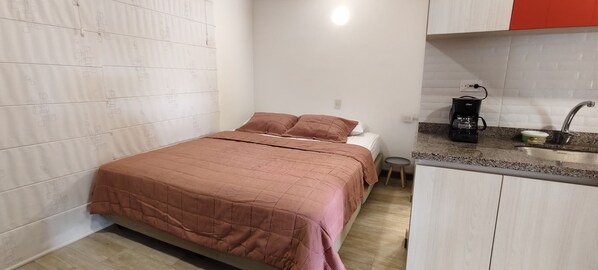 Un dormitorio con cama, un taburete pequeño y una kitchenette con cafetera.