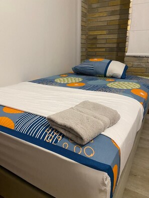 Una cama con una colcha azul y naranja estampada, una toalla gris doblada y un almohadón a rayas.