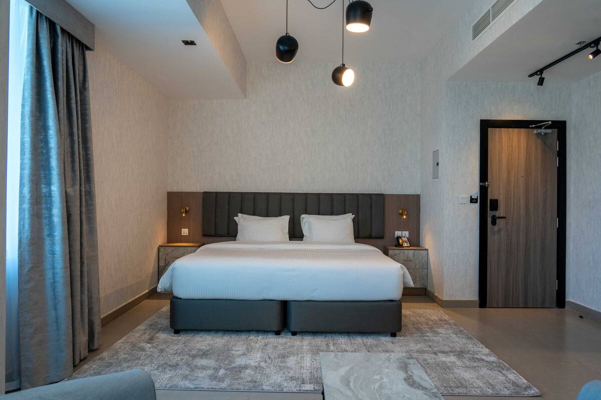 Habitación de hotel moderna con una cama grande, mesitas de noche, una puerta de madera y vistas al exterior.