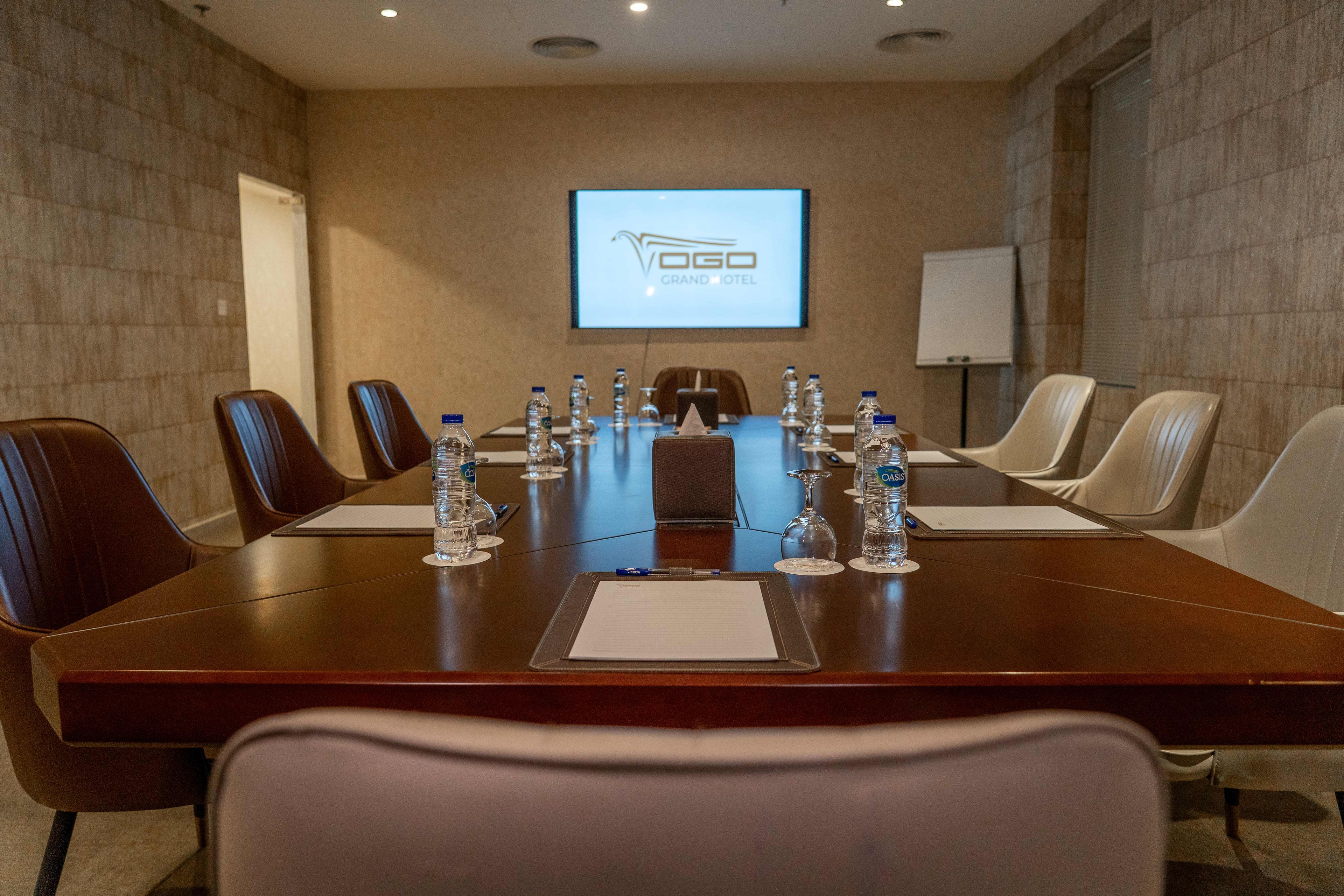 Uma sala de conferências com uma mesa comprida, cadeiras, garrafas de água e uma tela exibindo o nome do hotel.