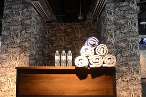 Una estantería de madera con botellas de agua y toallas enrolladas frente a una pared con textura.