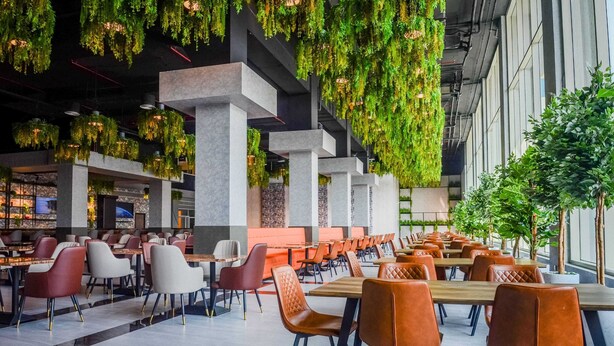 A modern dining area with tables and chairs, hanging greenery, and a view of the outdoors. - Vogo Grand Hotel (Dubai)