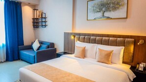 A hotel room with a bed, a sofa, a wall-mounted shelf with pictures, and a painting of a tree. - Vogo Grand Hotel (Dubai)