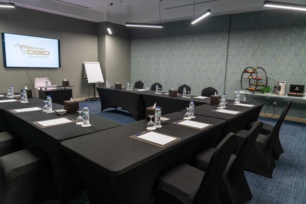 A conference room with long tables, chairs, water bottles, and a projector screen displaying 'VGO GRAND HOTEL'. - Vogo Grand Hotel (Dubai)