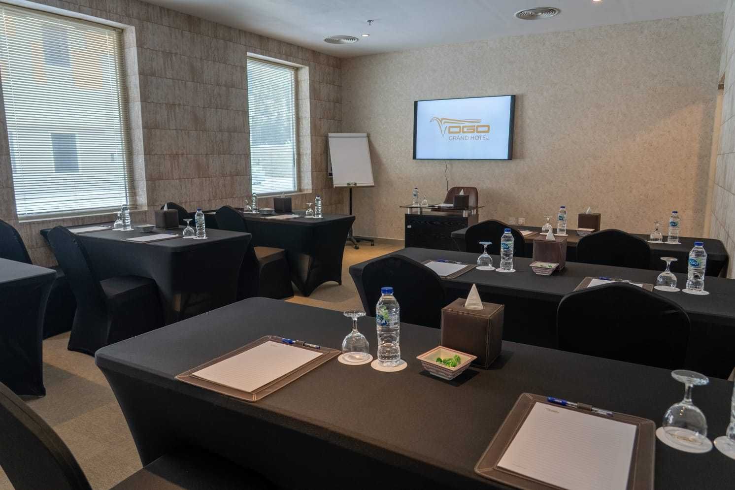 A conference room with tables, chairs, water bottles, and a screen displaying the Grand Hotel logo.