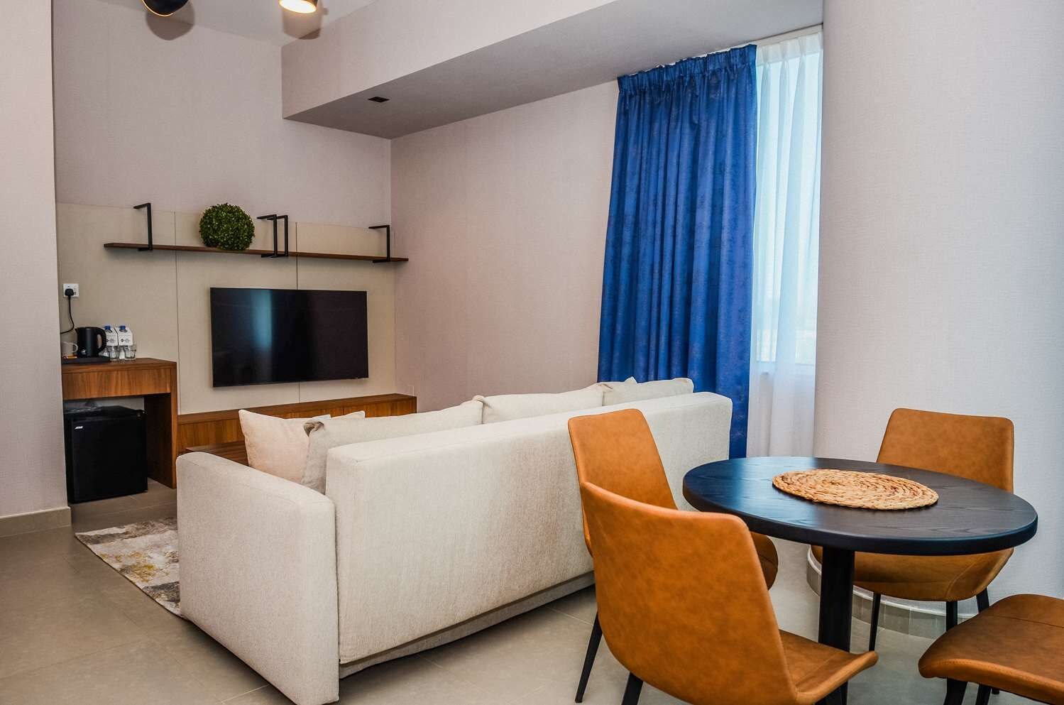 A modern living room with a sofa, a TV, a dining table with chairs, and a kitchen area with a refrigerator.