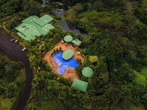 An aerial view of a resort with a central swimming pool, multiple green-roofed buildings, and lush surrounding vegetation. - Arenal Manoa Resort And Hot Springs (La Fortuna)
