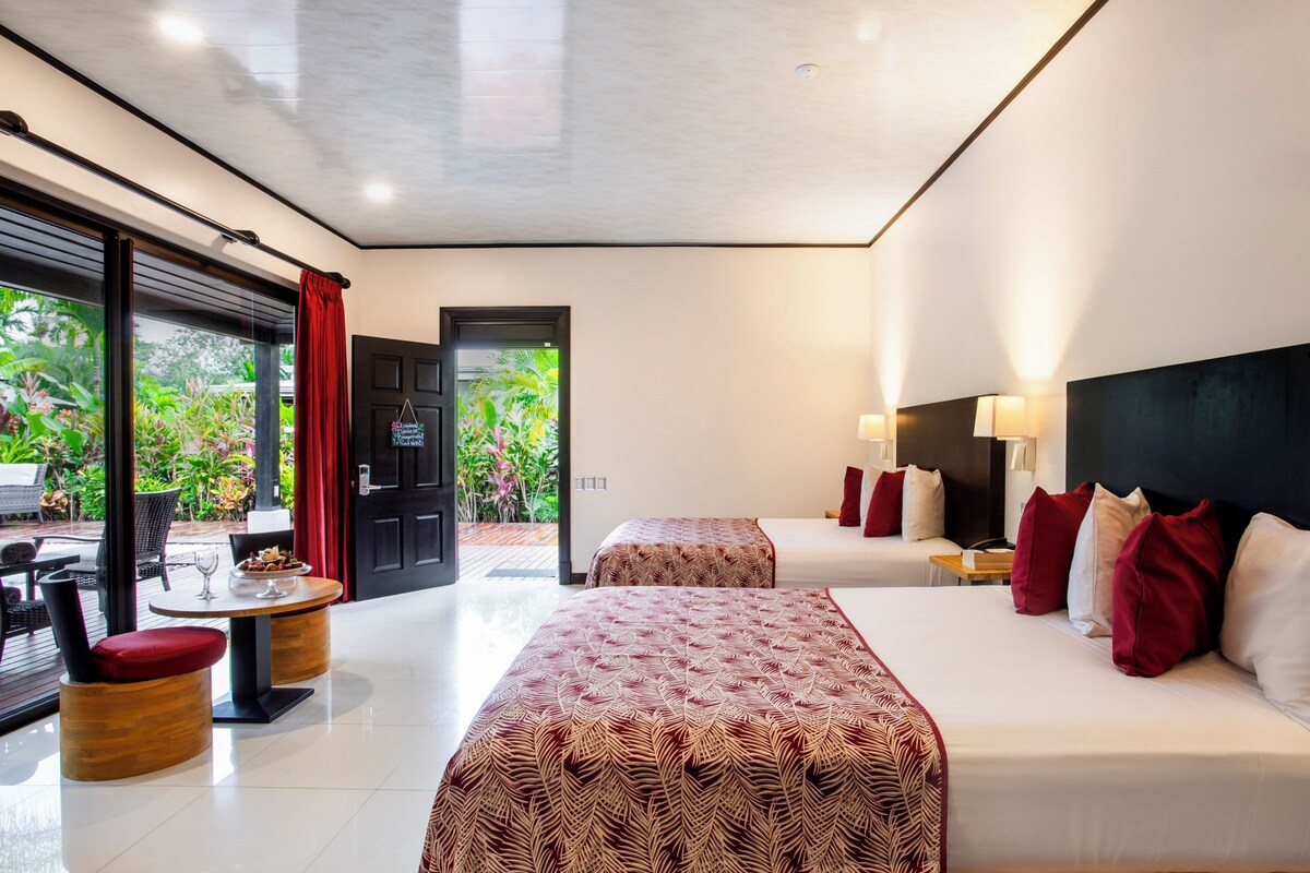A modern hotel room with two beds, a dining area, and a view of greenery outside.