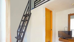 Una habitación con puerta de madera, escalera de metal negro, televisor en estante de madera y suelo de baldosas.