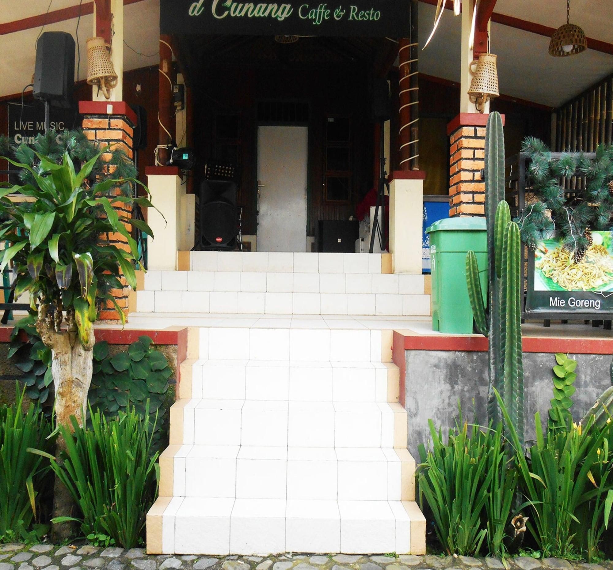 Entrada de um edifício com degraus que levam a uma porta, cercada por plantas e um letreiro do 'd Cunang Caffe & Resto'.