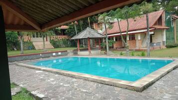Uma piscina com deck de pedra, um gazebo e uma casa com telhado vermelho ao fundo.
