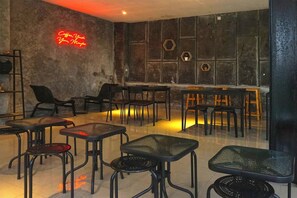 Un café con mesas y sillas dispuestas en un ambiente relajado, un letrero de neón en la pared y decoraciones hexagonales.