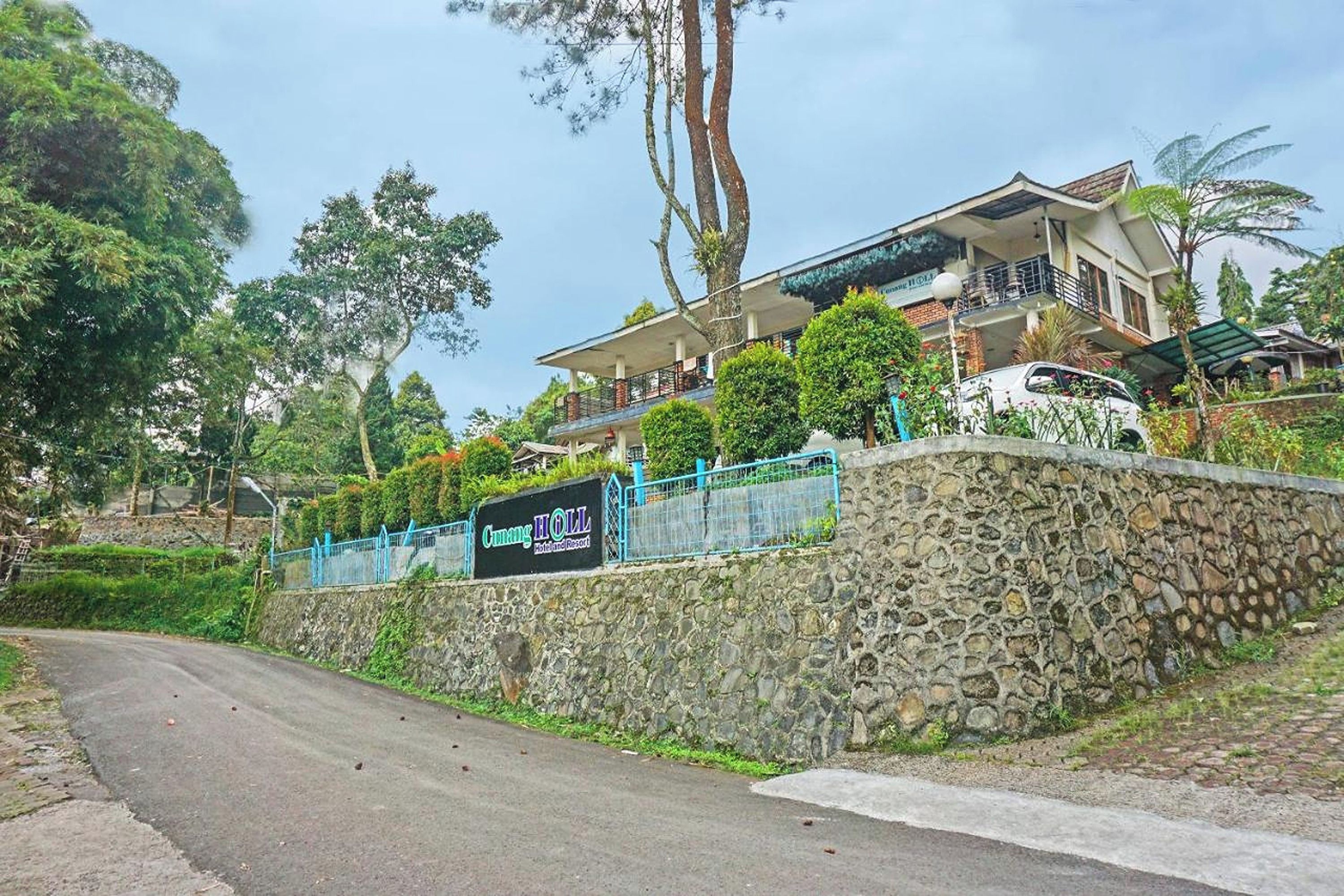 Um muro de pedra com uma placa indicando o hotel 'Cang HOLL', uma estrada sinuosa e uma árvore grande.