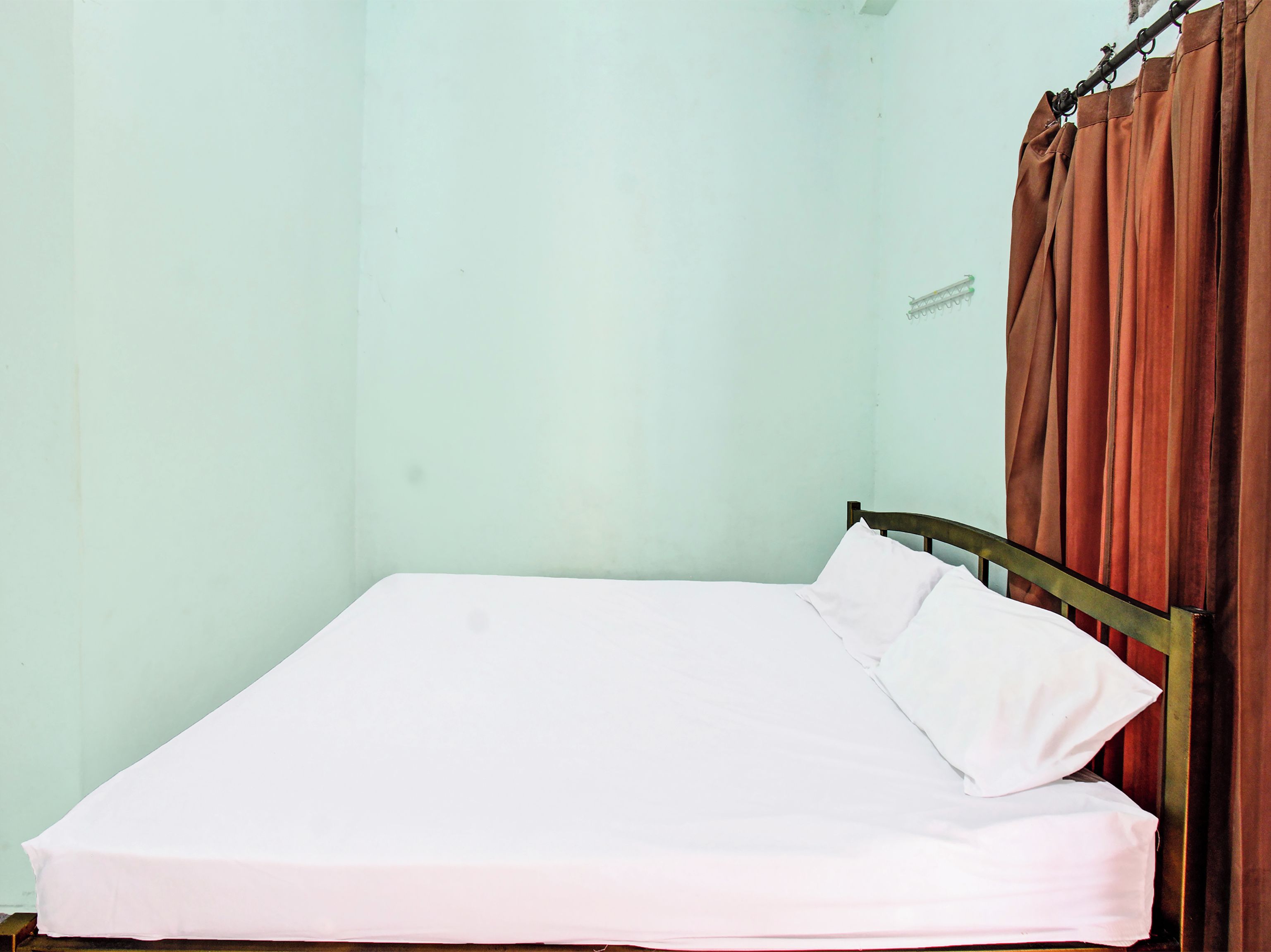 Un lit simple avec un cadre en métal et un matelas blanc, dans une pièce aux murs vert clair et dotée d’un rideau marron.