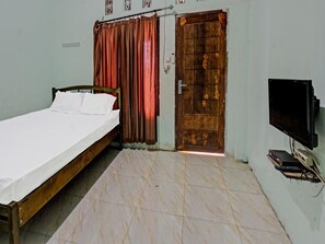 Una cama individual con ropa de cama blanca, una puerta de madera, un televisor montado en la pared y piso de baldosas.
