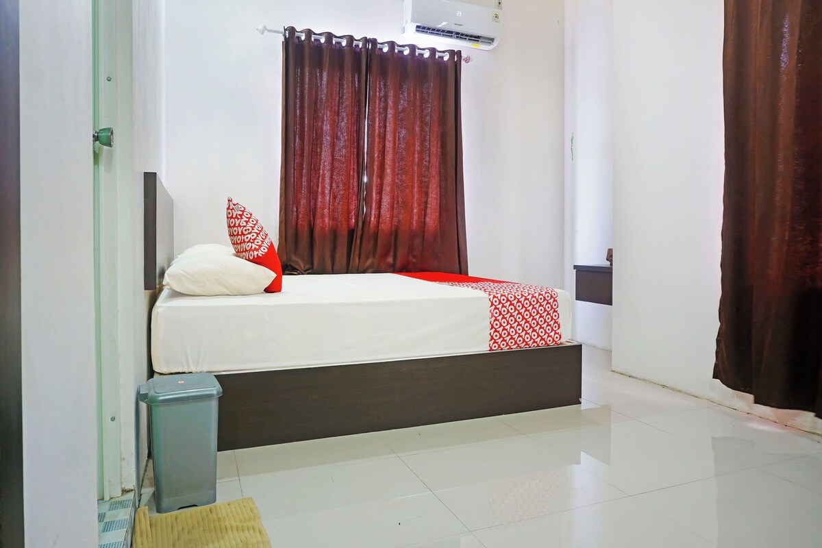 A bedroom with a bed, a window with curtains, an air conditioning unit, and a trash bin.