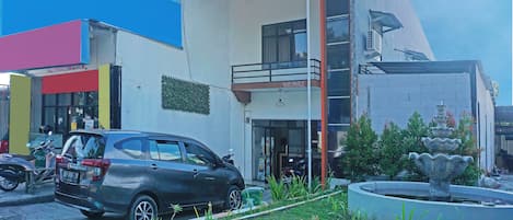 A modern building with a balcony, a car parked in front, and a small garden with a fountain.