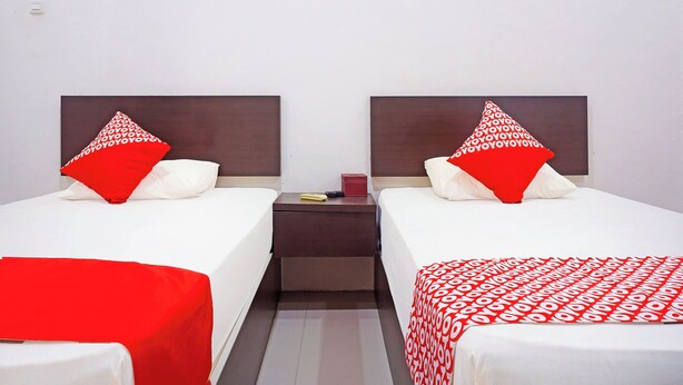 A hotel room with two beds, each with a red and white patterned pillow, a small bedside table with a book and a remote control, and a red blanket on one bed. - Oyo 91432 Dalle 5151 Jeneponto (Tanjung Pinang)