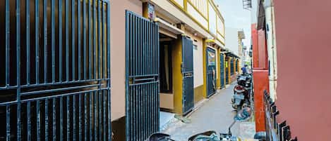 Uma rua estreita com edifícios em ambos os lados, uma motocicleta estacionada à direita e um portão à esquerda.