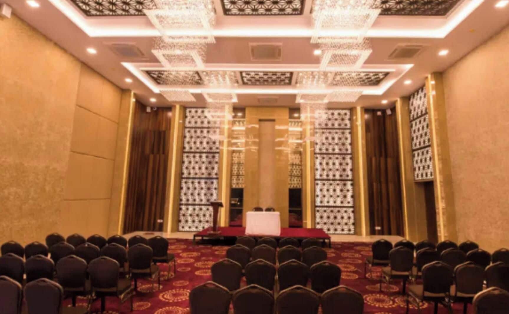 Une salle de conférence avec des rangées de chaises disposées face à une scène recouverte d’une nappe rouge.