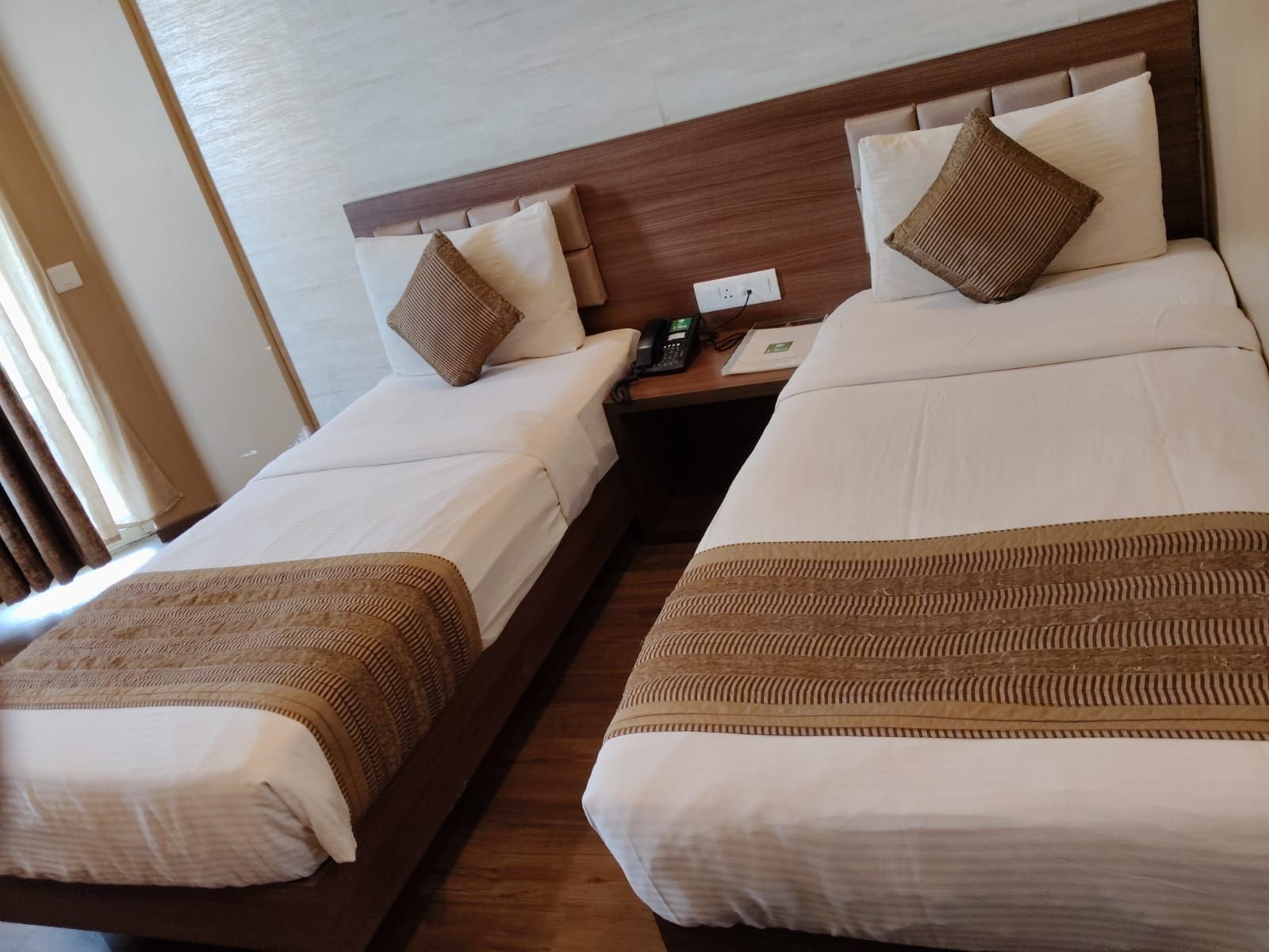 A hotel room with two single beds, a wooden headboard, a nightstand with a phone, and a window with curtains.