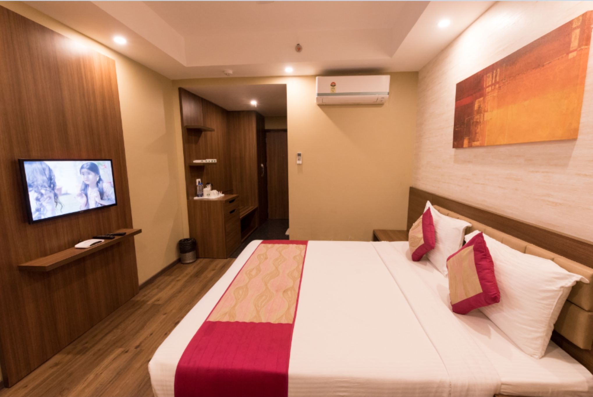 A hotel room with a bed, a TV mounted on the wall, a wooden floor, and an air conditioning unit.