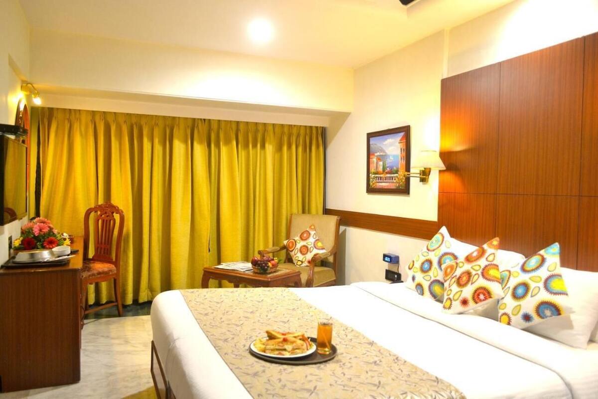 A hotel room with a bed, a desk with a lamp, a chair, a table with a fruit bowl, and a painting on the wall.