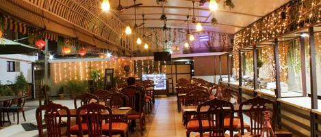 Un restaurante con mesas y sillas de madera, luces colgantes y un amplio ventanal con plantas.