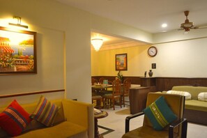 Une chambre d’hôtel avec un coin repas, un canapé agrémenté de coussins colorés, une horloge au mur et un ventilateur de plafond.
