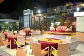 Un espace extérieur aménagé pour les repas, avec des chaises et des tables en osier, une scène avec des sièges et un espace couvert agrémenté de plantes.