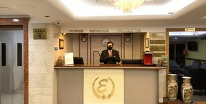 Un comptoir d’accueil d’hôtel avec un homme debout derrière, une horloge et un panneau indiquant « Caisse, réception, renseignements ».
