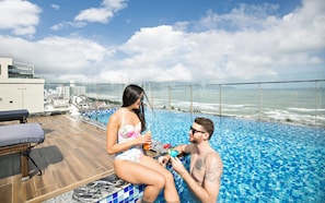 A couple relaxing by a pool with a city view, a lounge chair, and a glass railing. - Halina Hotel And Apartment (Da Nang)