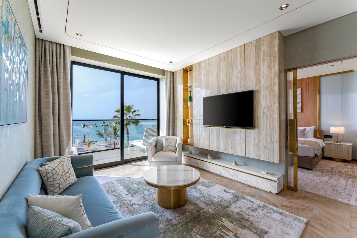 A modern living room with a large flat-screen TV, a sofa, a coffee table, and a view of the beach.
