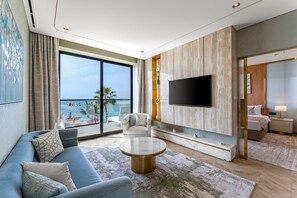 A modern living room with a large flat-screen TV, a sofa, a coffee table, and a view of the beach.