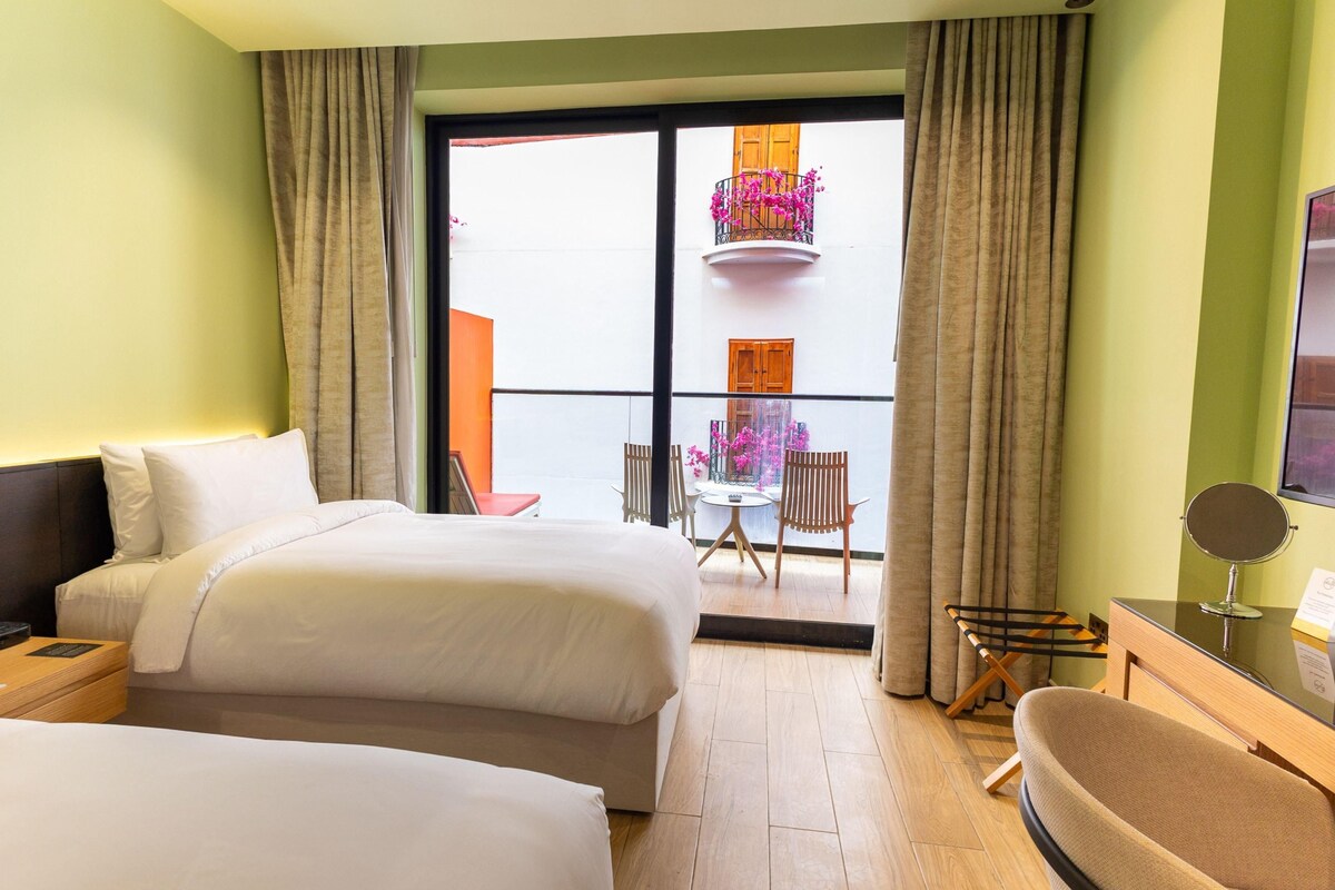 A hotel room with two beds, a small table, a chair, a TV, and a balcony with a view of the outside.