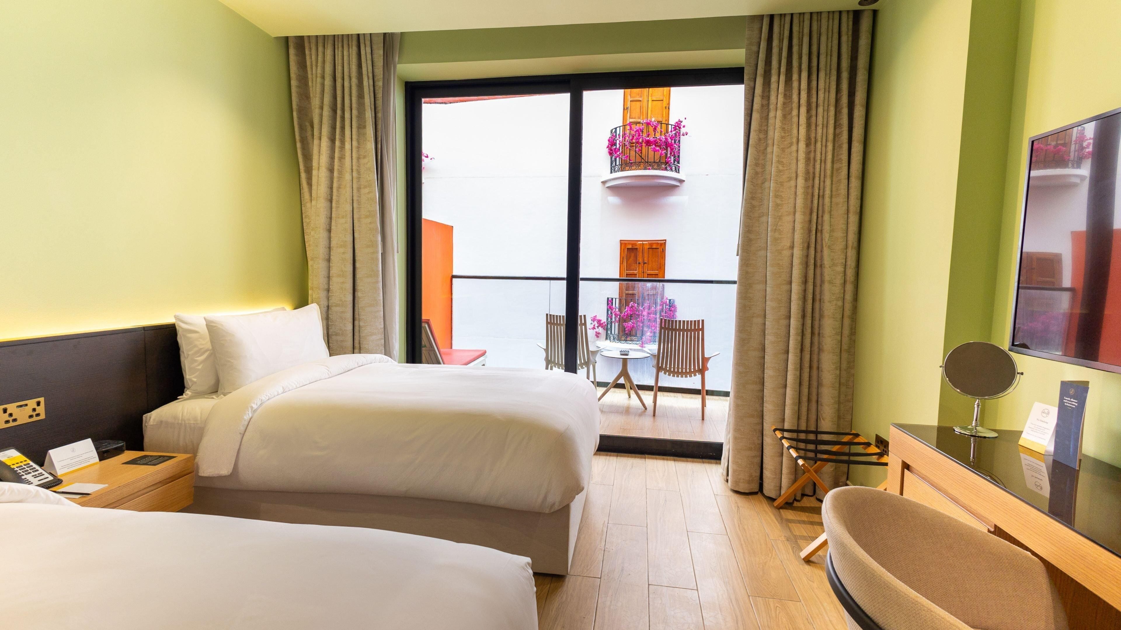 A hotel room with two beds, a small table, a chair, a TV, and a balcony with a view of the outside.