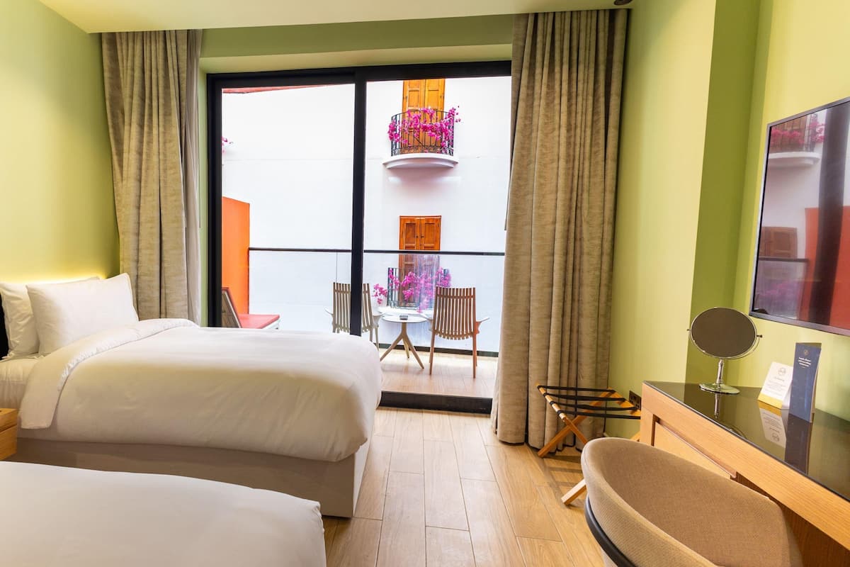 A hotel room with two beds, a small table, a chair, a TV, and a balcony with a view of the outside.