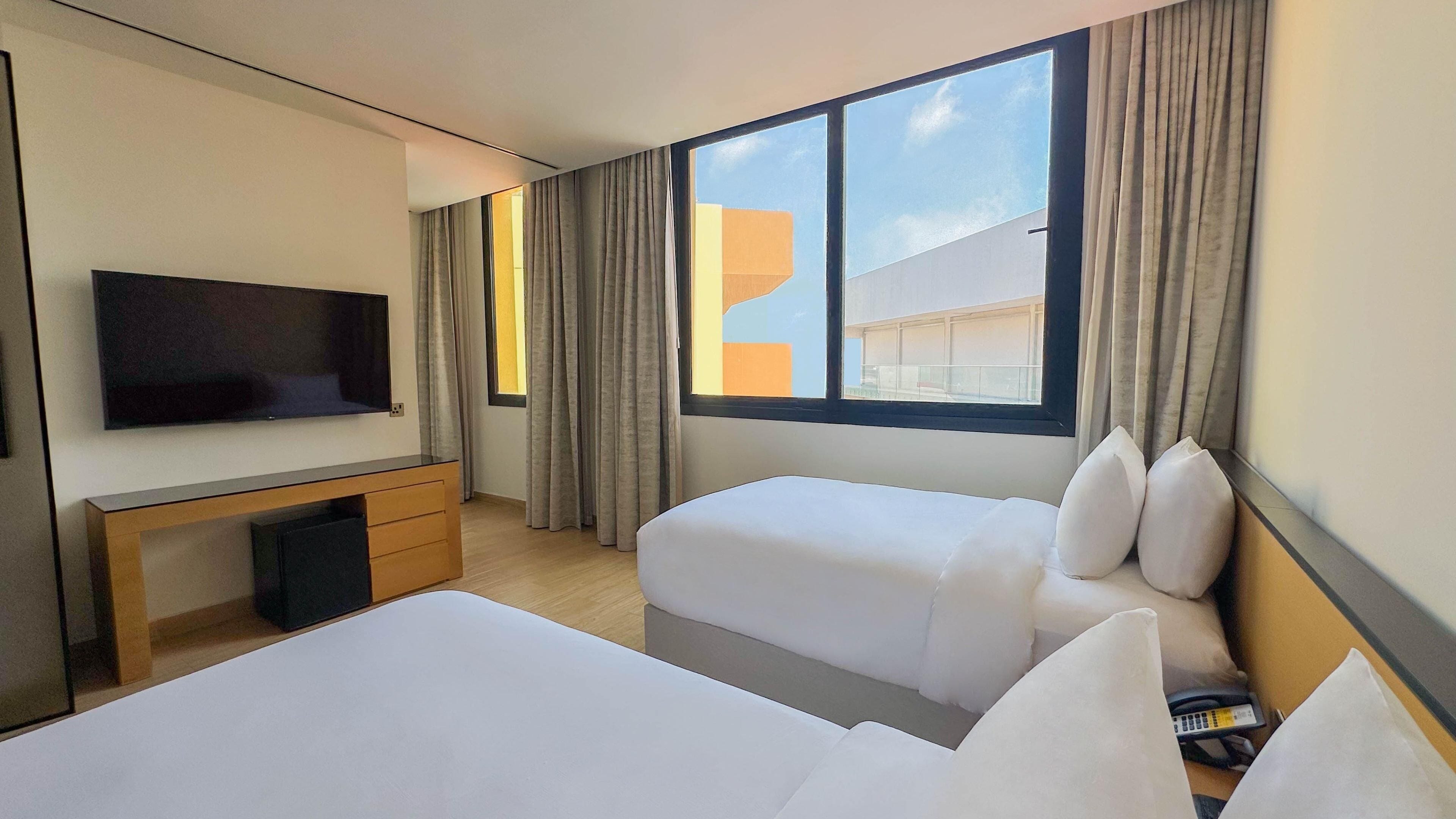 A hotel room with a TV, a desk, two beds, and a large window with curtains.