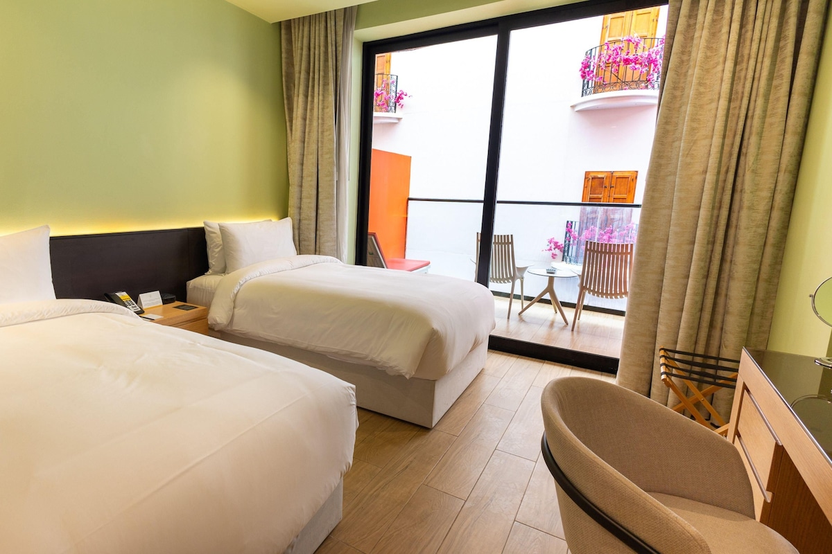 A hotel room with two beds, a small table, a chair, and a balcony with a view of a building and flowers.