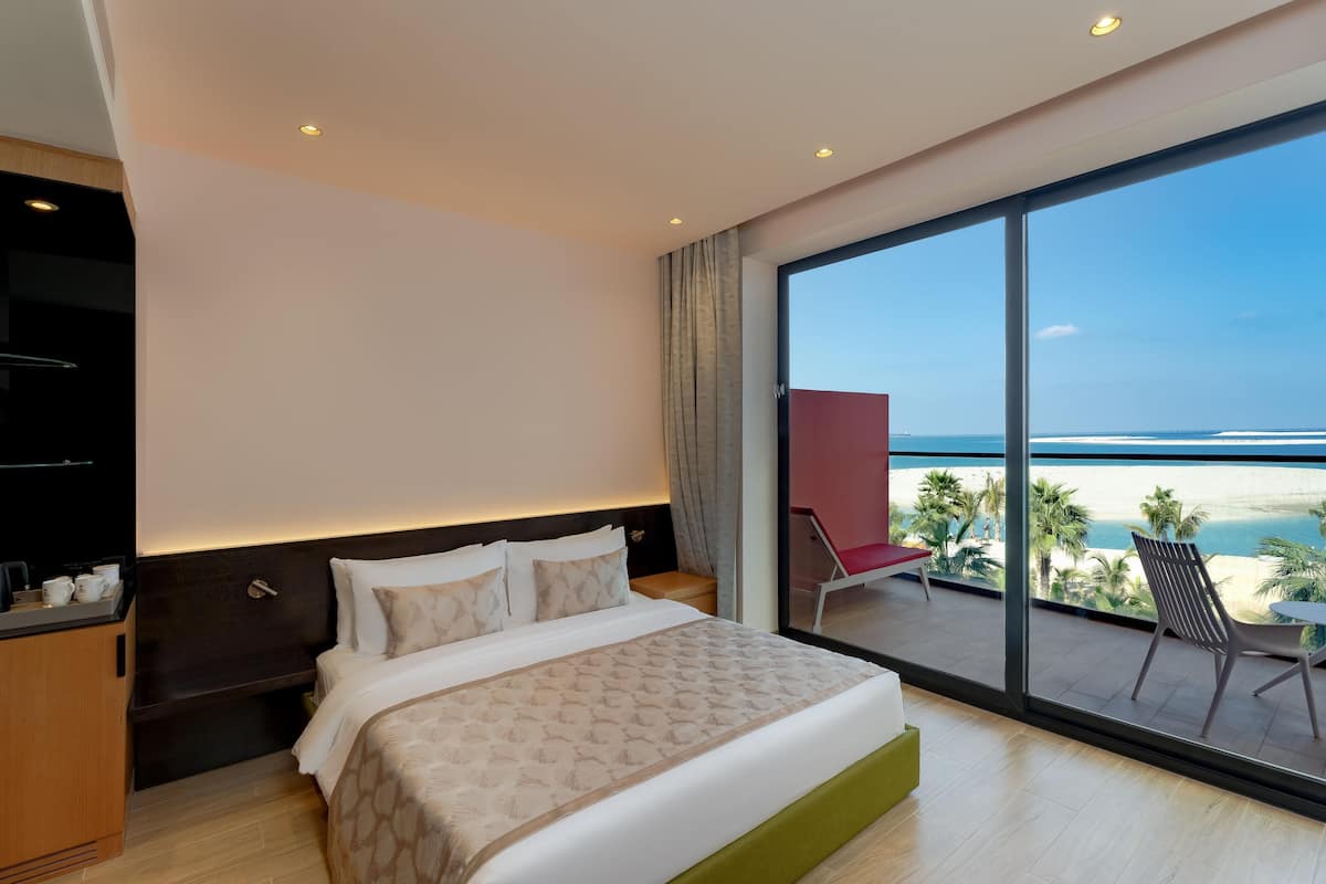 A modern hotel room with a large bed, a balcony with a view of the sea, and a seating area with a red chair.