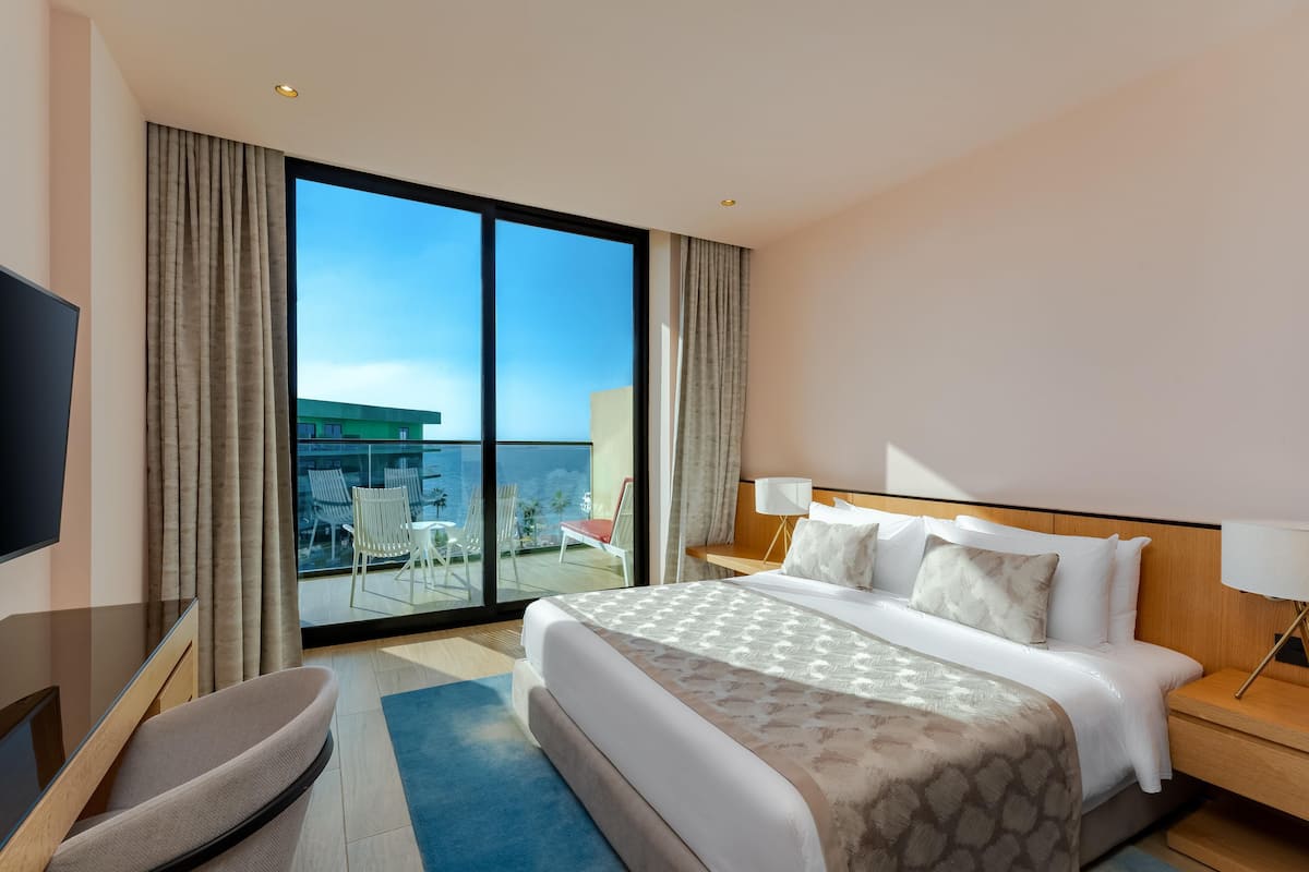 A hotel room with a large bed, a TV, a desk, and a balcony with chairs.