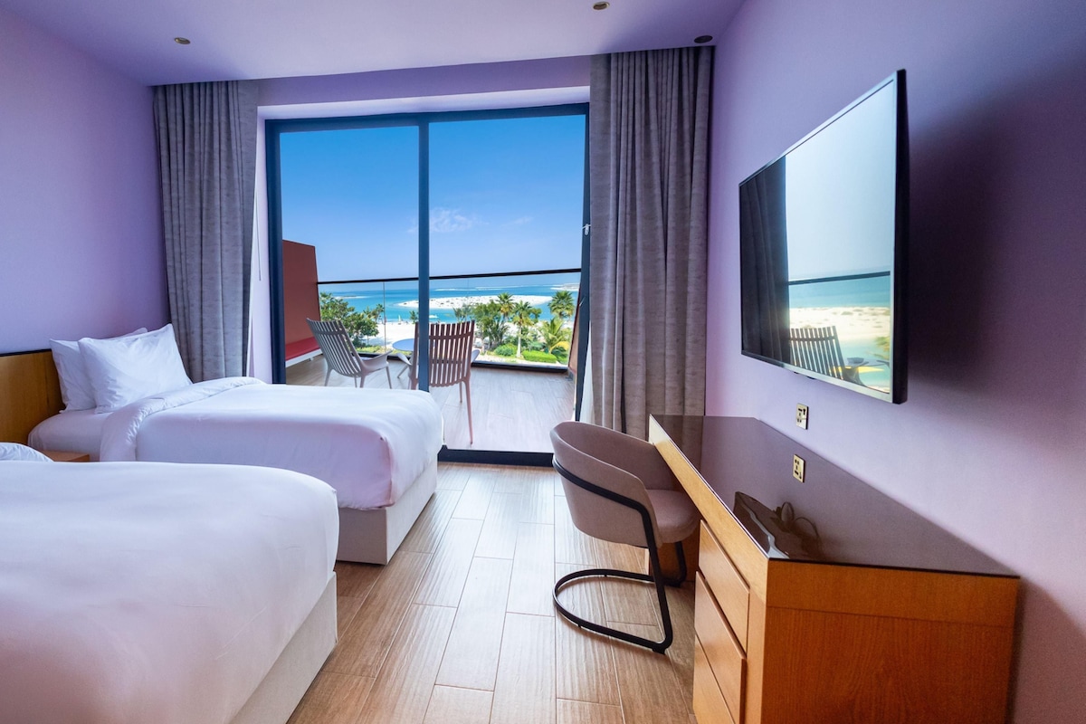 A hotel room with two beds, a desk, a chair, a TV, and a balcony with a view of the beach.