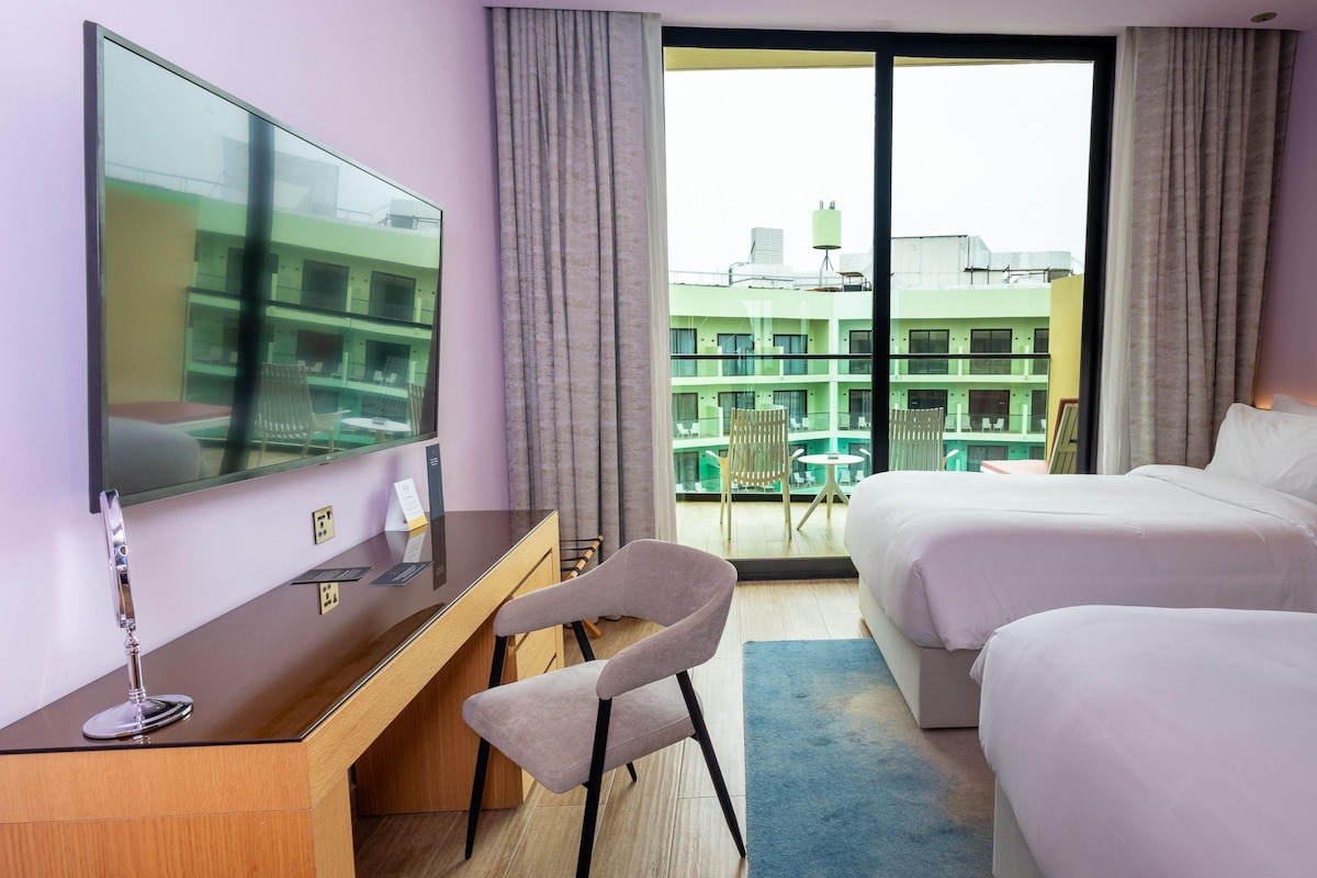 A hotel room with a desk, two beds, a TV, and a balcony with a city view.