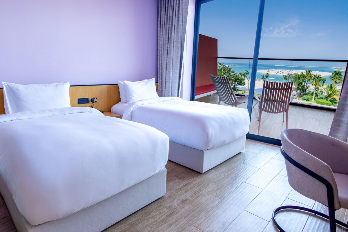 A hotel room with two beds, a balcony with a view of the beach, and a chair.