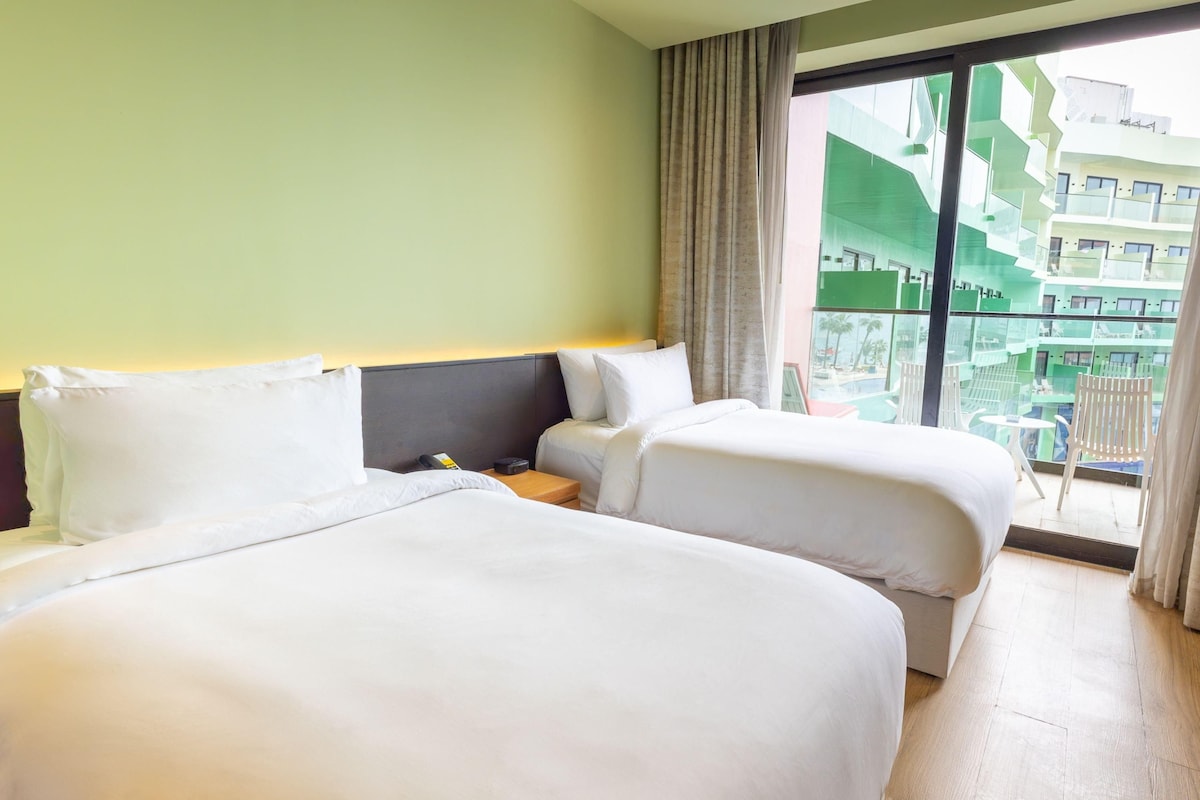 A hotel room with two beds, a nightstand, a phone, and a view of a green building through the window.