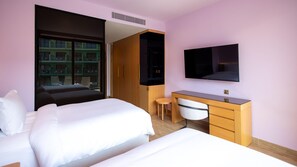 Hotelkamer met twee bedden, een bureau, een stoel, een televisie en een groot raam met uitzicht op gebouwen.