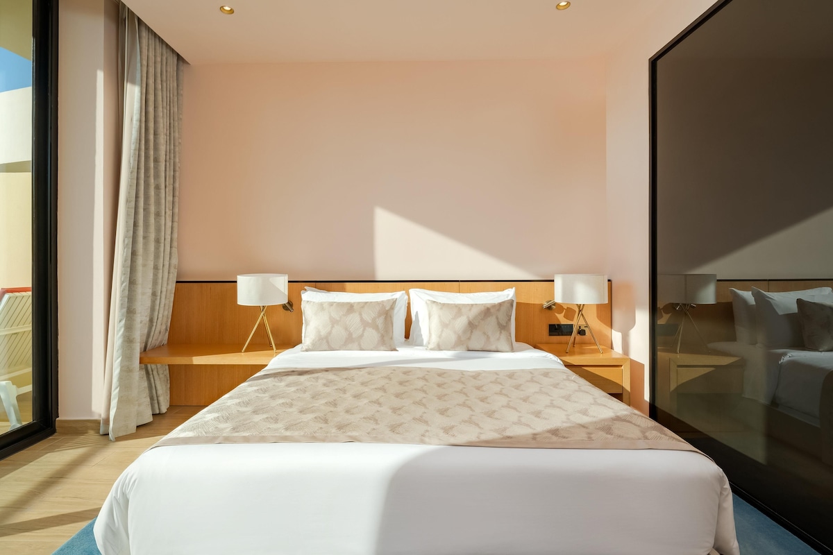 A hotel room with a large bed, two bedside tables, a window with curtains, and a view of the outside.